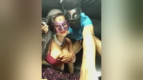 Video of a woman with long black hair and purple glasses, wearing a revealing red top, and a man with a black mask and blue shirt, both in a car.
