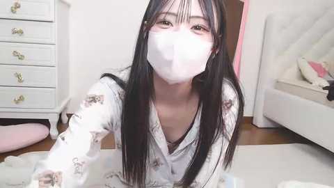 Video of an Asian woman with long black hair and a white mask, wearing a floral-patterned robe, sitting on the floor in a bright, tidy bedroom with white furniture.