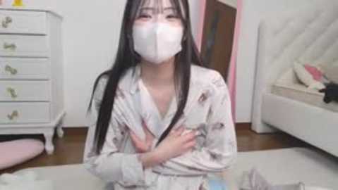 Video of an East Asian woman with long black hair, wearing a white mask, floral pajamas, and sitting on a rug in a white bedroom with a white dresser, bed, and pink chair.