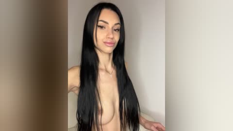 Video of a topless transgender woman with long, straight black hair, fair skin, and small breasts, posing confidently indoors with a plain white wall background.