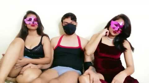 Video of three masked individuals seated against a plain white wall; two women in black and red outfits, and a man in a black tank top and blue shorts.
