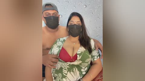Video of a topless man with a black mask and cap, and a woman wearing a green floral shirt and red bra, both indoors with a textured wall background.