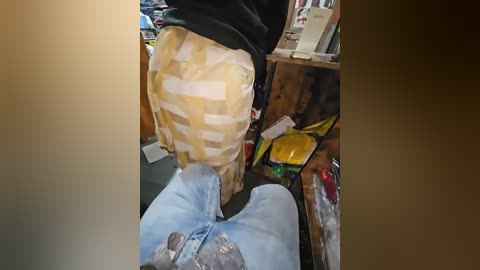 A video shows a person's legs wearing light blue jeans and a yellow plaid shirt, sitting on a cluttered wooden shelf with books, papers, and a yellow package.