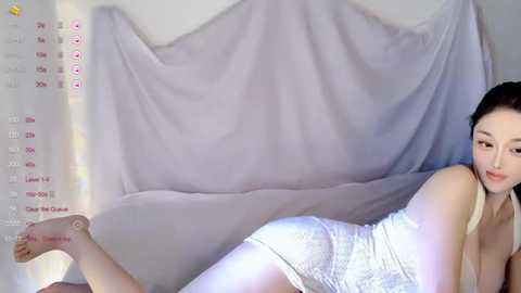 A video of a fair-skinned Asian woman with black hair, lying on a bed, wearing white lace panties, with a white fabric draped behind.