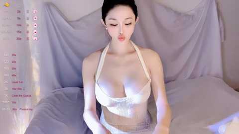 A video of a young East Asian woman with fair skin, dark hair, and a slender physique, wearing a revealing white halter top that accentuates her large breasts. She sits on a bed draped in light purple sheets, with a soft, ambient lighting.