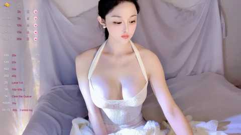 A video of an East Asian woman with fair skin, long black hair, and large breasts in a white halter dress, sitting on a bed draped with white sheets, with a soft, romantic ambiance.