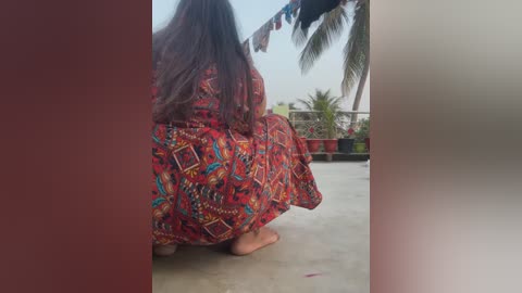 A video captures a woman with long, dark hair in a colorful, patterned sari squatting on a concrete balcony. She faces a palm tree and a potted plant. The background shows a clear blue sky.