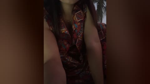 A video of a woman with medium skin tone wearing a sleeveless, patterned red dress, her face partially visible. The background is dimly lit, featuring blurred objects, possibly indoor furniture.