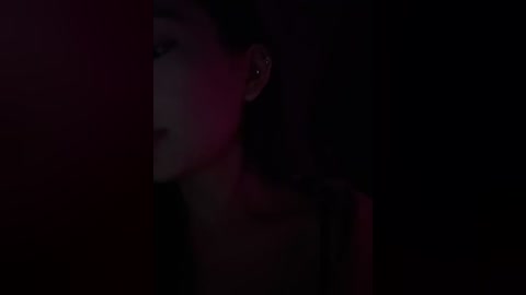 Video of a woman's face, partially lit with dark shadows, wearing heavy makeup, including dark eyeshadow, red lipstick, and subtle earrings.