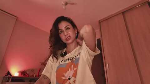 Video of a young woman with long, wavy dark hair, wearing a white t-shirt with a cartoon character, posing in a dimly lit room with wooden wardrobes and a red lamp.