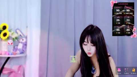 Video of a young Asian woman with long black hair, wearing a black dress, drinking green liquid, in a pastel-toned room with a flower vase, and a live-streaming screen showing stats.