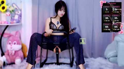 Video of an Asian woman in black lingerie, sitting with legs spread, watching a phone. Background features a plush toy, a clock, and a digital game screen.
