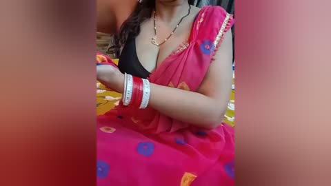 Video of a woman in a red sari with blue polka dots, wearing a black bra and multiple bangles, sitting on a bed with yellow sheets, in a blurry, intimate indoor setting.
