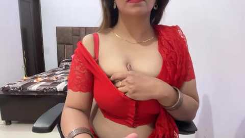 Video of a curvy woman with medium skin tone, wearing a red lace bralette and matching shawl, sitting on a black chair in a bedroom with a bed and dark wood headboard.