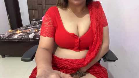 Video of a South Asian woman with medium skin tone, wearing a red lace bra and matching skirt, seated on a black chair in a simple room with a bed and brown door.