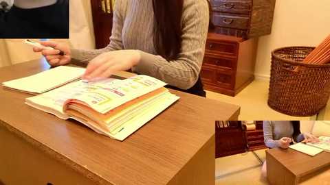 Video of an Asian woman in a gray sweater, seated at a wooden desk, flipping through a colorful book, with a wicker basket and wooden furniture in the background.