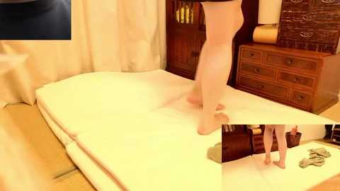 Video of a woman in a beige nightgown standing on a white bed, with a wooden nightstand and brick wall in the background.