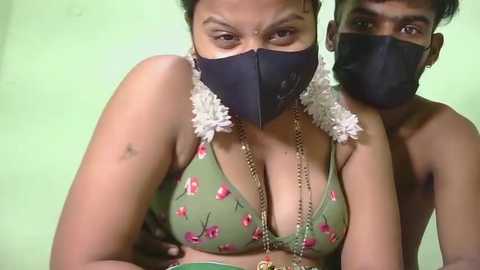 Video of a topless, dark-skinned woman wearing a green floral bra with a black face mask, and a man behind her with a similar mask, both in a dimly lit room with a green wall.