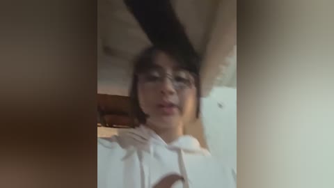 A blurry, out-of-focus video of a young person with dark hair and glasses, wearing a white hoodie, standing indoors with a wooden ceiling and blurred background.