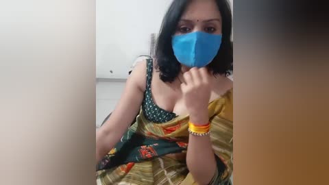 A video shows a South Asian woman with medium skin tone, wearing a blue mask, green sari with floral patterns, and yellow bangles, sitting indoors.
