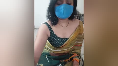 A video of a South Asian woman with medium skin tone, wearing a blue mask, black hair, and a yellow-green sari with a green blouse. She's seated indoors with a blurred background.
