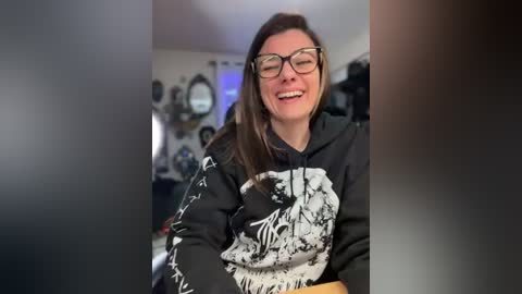 Video of a smiling, fair-skinned woman with glasses, wearing a black graphic hoodie, sitting indoors, with a blurred background of a cozy room.