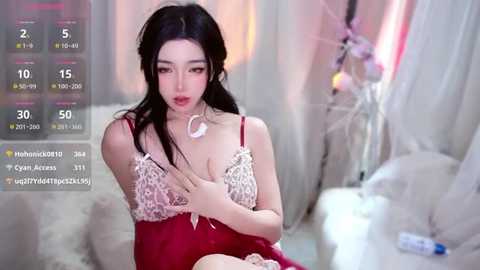 A video of an East Asian woman with long black hair, wearing a revealing red dress with lace, sitting in a plush, white room.
