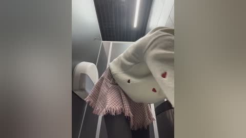 A video of a woman in a white coat and red checkered skirt, standing in a modern, minimalist bathroom with a black and white tiled floor and a circular mirror.