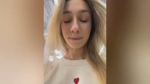 Video of a young Caucasian woman with light blonde hair, wearing a white sweatshirt with a red heart graphic, eyes closed, and a serene expression.