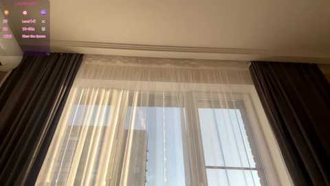 Video of a modern hotel room with a large window covered by sheer white curtains and dark, heavy drapes. The ceiling is painted a light beige, and a TV screen displays a channel guide.