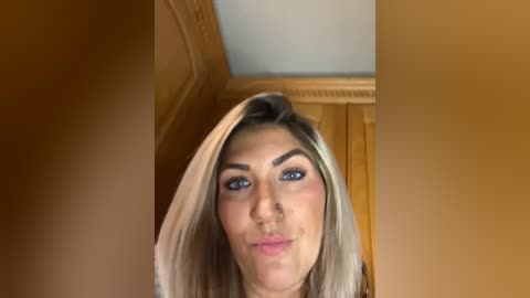 A video of a young woman with light brown hair and blue eyes, standing indoors against a warm-toned, wooden wall with a ceiling light. She has a subtle, confident smile.