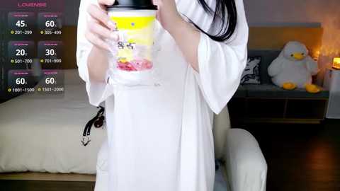 Video of a person wearing a white bathrobe, holding a yellow and white container labeled \"Soy Milk.\" Background features a plush duck, a bed, and a lamp.