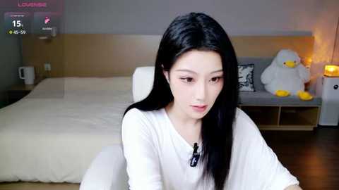 Video of a young Asian woman with long black hair, wearing a white shirt, sitting in a modern bedroom with a bed, stuffed animals, and a warm light.