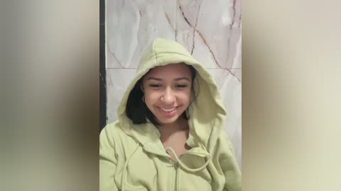 A video of a young woman with medium brown skin, medium build, and shoulder-length dark hair, smiling while wearing a light green hoodie in a bathroom with marble tiles.
