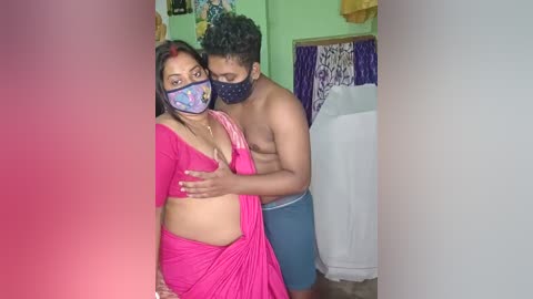 Video of a topless man in blue shorts embracing a woman in a pink sari with a colorful face mask, in a dimly lit room with green walls.