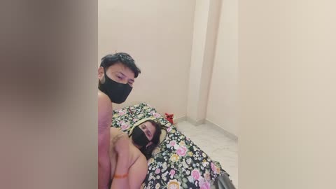A video shows a couple in a dimly lit room, both wearing black masks. The man, topless, stands behind the woman, who has shoulder-length brown hair and is partially nude, lying on a bed with a floral-patterned sheet.
