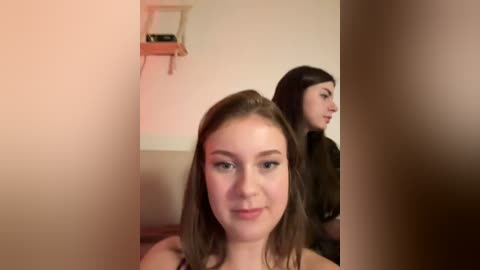 Video of two young women with light skin, one in the foreground with shoulder-length brown hair, the other in the background with shoulder-length black hair. They stand in a simple room with beige walls and a wooden shelf.