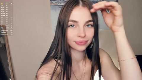 Video of a young Caucasian woman with long, straight, dark brown hair, fair skin, and blue eyes, wearing a white tank top. She is indoors, adjusting her hair with her right hand, against a beige wall with a framed photo behind her.