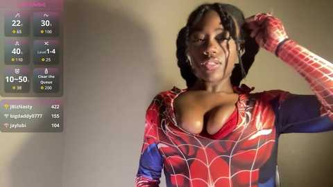 Video of a dark-skinned woman with large breasts wearing a tight Spider-Man costume, adjusting her hair against a beige wall.