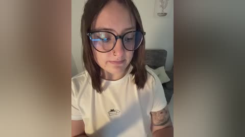 Video of a young woman with fair skin, shoulder-length brown hair, and large glasses. She wears a white T-shirt with a small logo. Background shows a beige wall and a gray couch.
