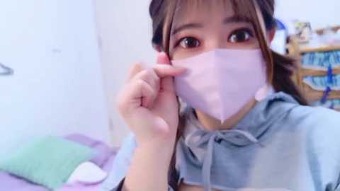 Video of an Asian woman with brown hair, wearing a pink surgical mask and light gray hoodie, sitting on a bed with pastel pillows in a white room.