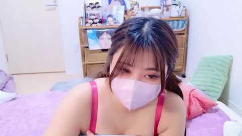 Video of an East Asian woman in a pink bra and face mask, lying on a bed with pastel pillows, in a brightly lit room with a bookshelf and white walls.