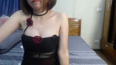 Video of a light-skinned woman with a short bob haircut, wearing a black strapless dress with red rose appliqu\u00e9s and a choker necklace, sitting on a bed in a simple, wooden-furnished room.