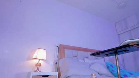 Video of a minimalist, purple-lit bedroom with a neatly made bed, white bedding, a wooden headboard, and a small bedside table with a lamp.