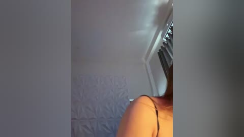 Media: A video of a person's back, wearing a sleeveless top, standing in a dimly lit room with white textured wallpaper and a white bedsheet. The image is slightly blurred and out of focus.