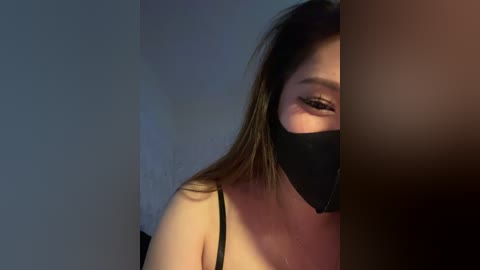 Video of an Asian woman with long black hair, partially obscured by a black face mask, wearing a black spaghetti strap top. She has light skin and is looking to the side.