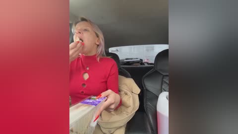 Video of an elderly woman with short, light blonde hair, wearing a red sweater, eating a sandwich in a car, holding a bag of chips, with beige jacket draped over seat.