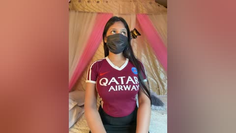 Video of a young woman with medium brown skin, wearing a black face mask, burgundy \"Qatar Airways\" jersey, and black pants, sitting in a bed with beige curtains and pink drapes.