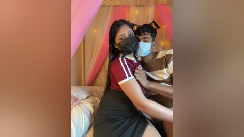 Video of an Indian couple in a cozy, dimly lit bedroom; woman with black hair, red shirt, black skirt, and face mask; man in striped shirt, face mask, embracing her.