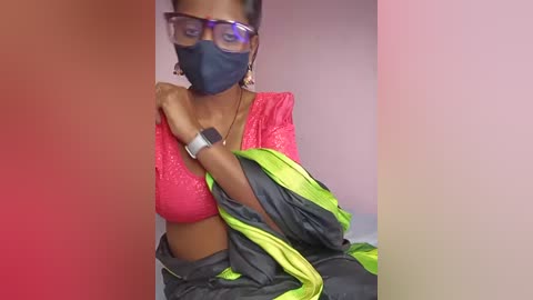 A video shows a woman in a bright pink blouse with a black face mask and black-framed glasses, holding a black and green sari. She has a medium-dark skin tone.